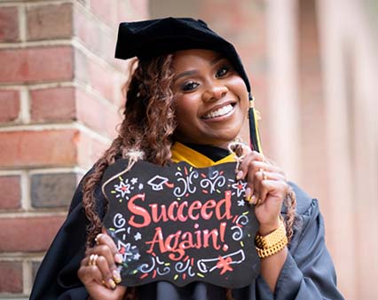 A female student celebrating with graduation cap and gown holding a sign that says succeed again.